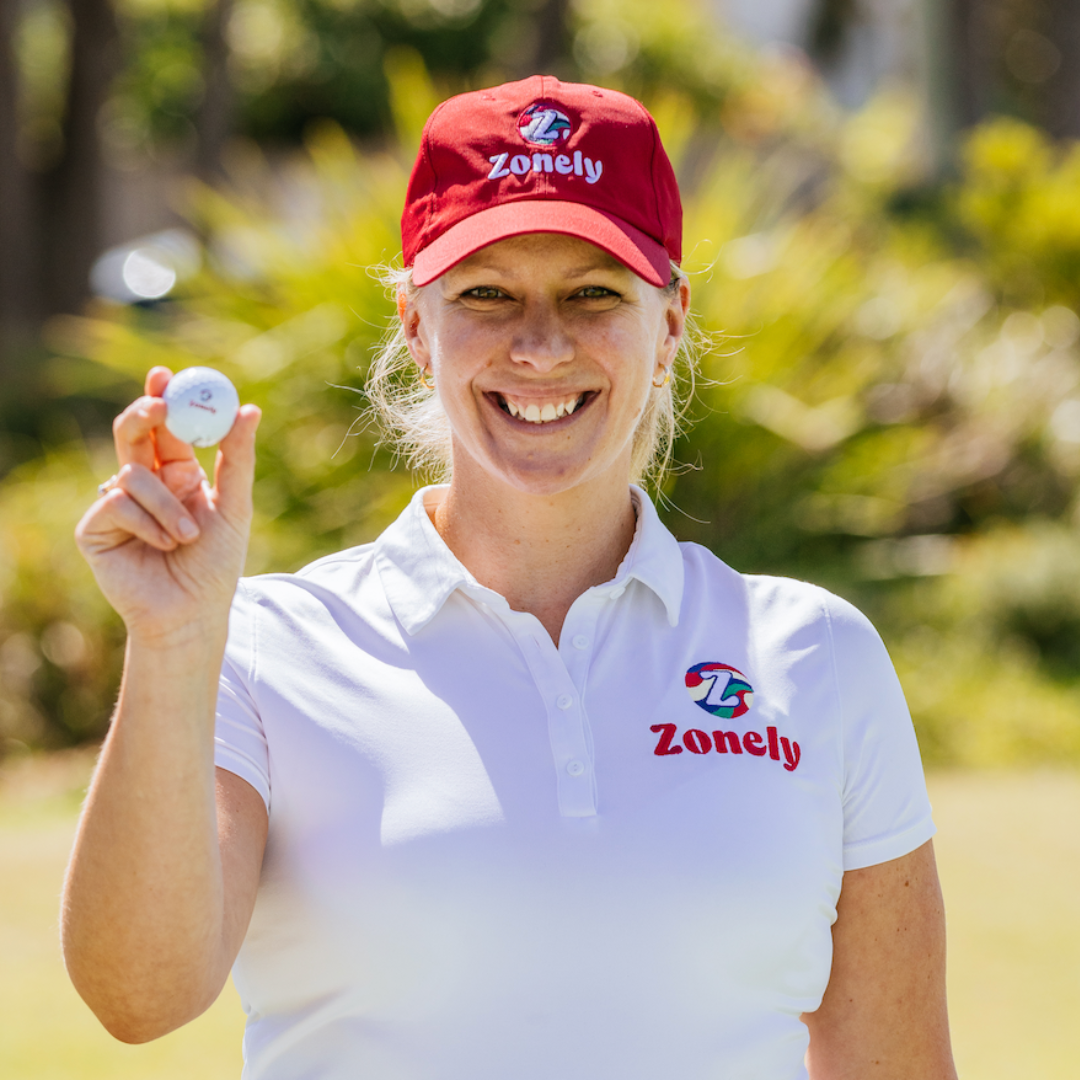 A smiling woman in a Zonely cap and shirt holds up a golf ball outdoors.